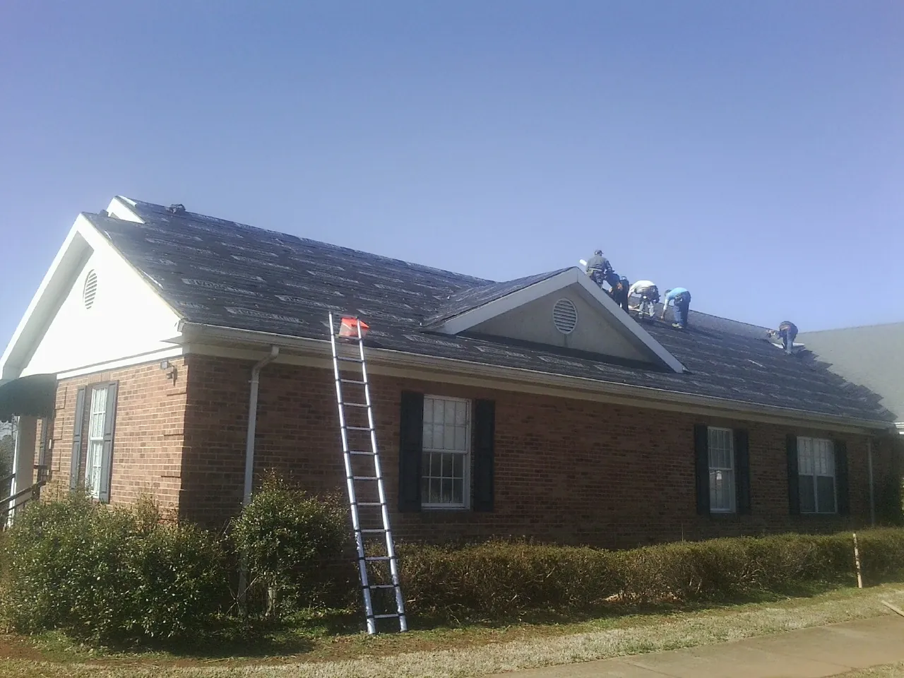 Crew tearing shingles off a brick ranch roof with a ladder set — Metal Roof Repair work by our crew in Fort Edward