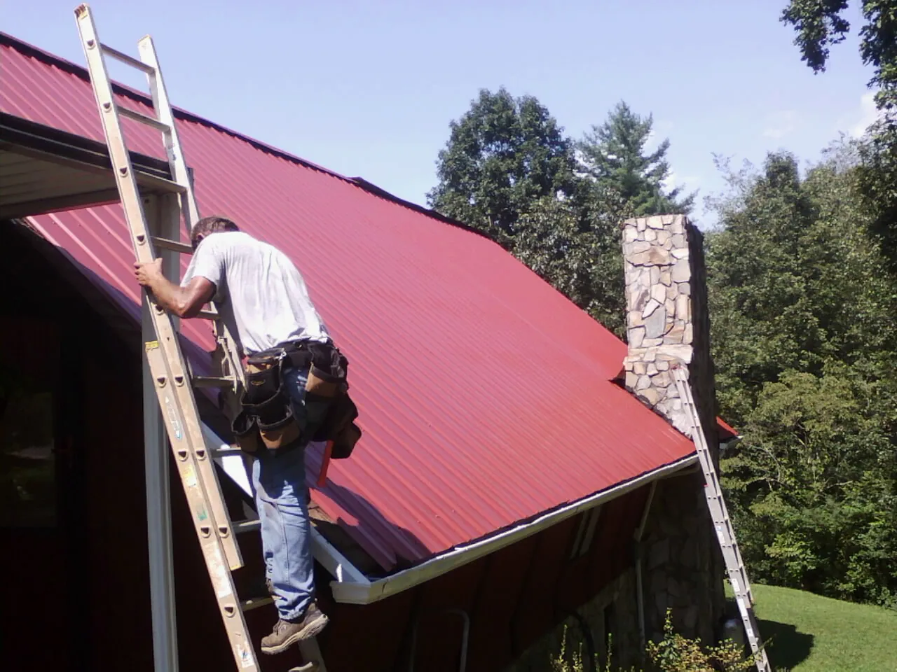 Roofer on a ladder beside a red metal roof and stone chimney — Soffit & Fascia Repair by Roofing Brothers in Fort Edward